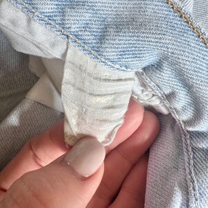 May include: Close-up of light blue denim jeans. The image focuses on the interior waistband and a small, partially visible tag. The denim has a classic, faded appearance, with visible stitching and a textured surface. The tag appears to be made of a light-colored fabric.