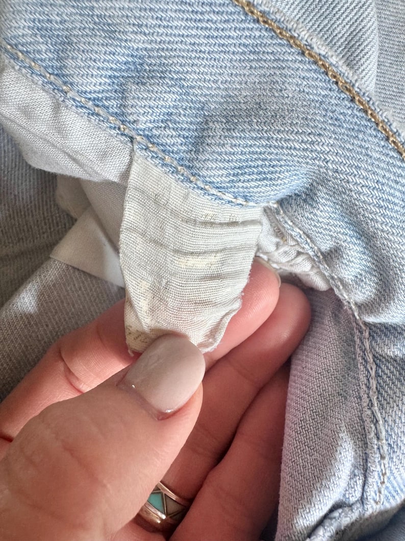 May include: Close-up of light blue denim jeans. The image focuses on the interior waistband and a small, partially visible tag. The denim has a classic, faded appearance, with visible stitching and a textured surface. The tag appears to be made of a light-colored fabric.
