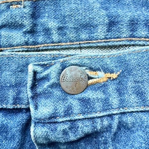 May include: Close-up of blue denim jeans, showcasing the button and buttonhole. The button is a tarnished metal with the word "Wrangler" embossed on it. The stitching is a contrasting gold color, highlighting the details of the jeans.
