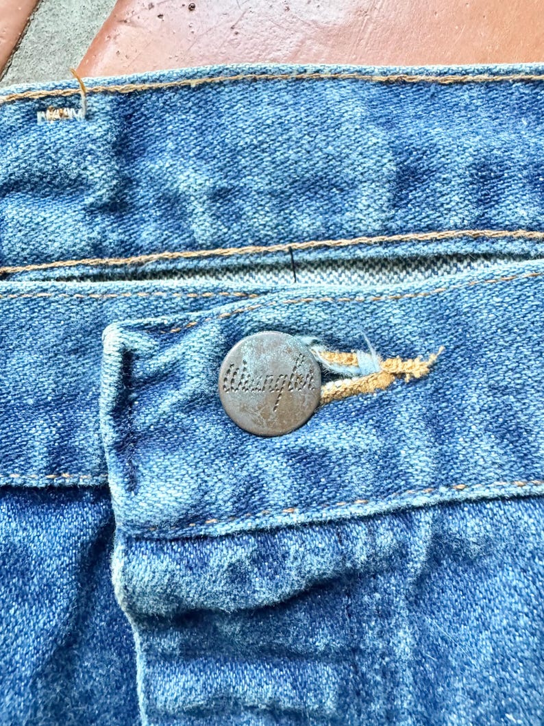 May include: Close-up of blue denim jeans, showcasing the button and buttonhole. The button is a tarnished metal with the word "Wrangler" embossed on it. The stitching is a contrasting gold color, highlighting the details of the jeans.