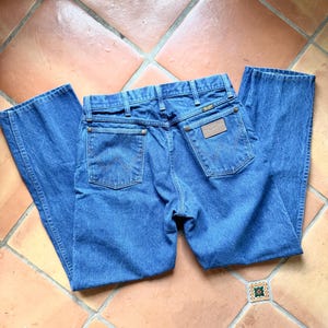 May include: Blue denim jeans with a classic five-pocket design. The jeans feature a Wrangler brand patch on the back and are laid flat on a tiled surface. The jeans have a straight leg cut.