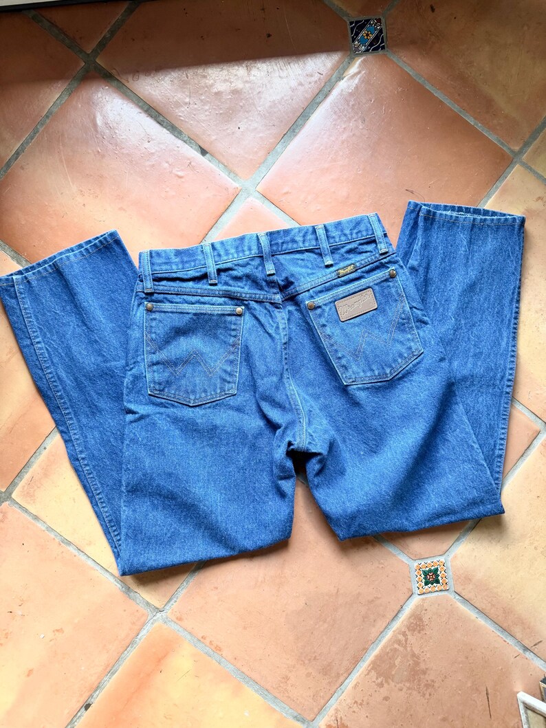 May include: Blue denim jeans with a classic five-pocket design. The jeans feature a Wrangler brand patch on the back and are laid flat on a tiled surface. The jeans have a straight leg cut.