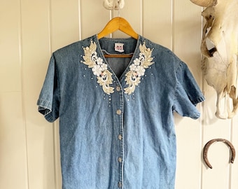 May include: A blue denim button-up shirt with short sleeves and a floral embroidered collar. The embroidery is in shades of white, gold, and pink.