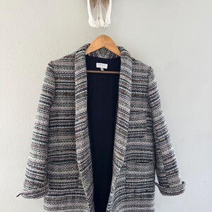 May include: A multi-colored tweed blazer with a black interior hangs on a wooden hanger. The blazer features a mix of green, brown, and gray threads. The sleeves are rolled up, and the blazer is open, revealing the black lining.