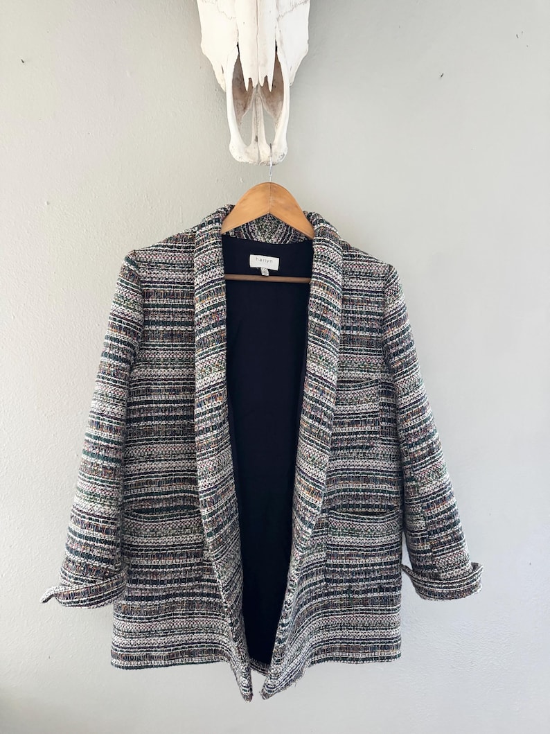 May include: A multi-colored tweed blazer with a black interior hangs on a wooden hanger. The blazer features a mix of green, brown, and gray threads. The sleeves are rolled up, and the blazer is open, revealing the black lining.