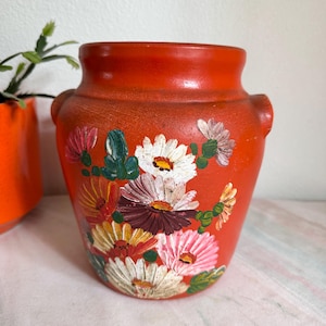 May include: Orange ceramic planter with hand-painted floral design. The flowers are white, pink, yellow, and burgundy.