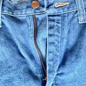 May include: Close-up of blue denim jeans with a visible zipper and a button closure. The button is a copper color and has the word "Wrangler" engraved on it. The jeans are slightly faded, showing signs of wear.