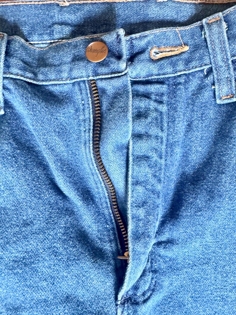 May include: Close-up of blue denim jeans with a visible zipper and a button closure. The button is a copper color and has the word "Wrangler" engraved on it. The jeans are slightly faded, showing signs of wear.