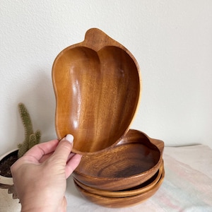 May include: Set of wooden mango-shaped bowls. The bowls are made of a warm-toned wood and have a smooth, polished finish. The set includes a larger bowl and several smaller, round bowls. The bowls are stacked on top of each other.