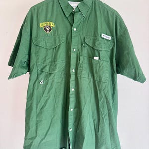 May include: A short-sleeved green button-down shirt featuring a Baylor bear logo patch on the right chest and a Columbia brand label on the left chest pocket. The shirt includes two front chest pockets with flaps and a button-up front closure.