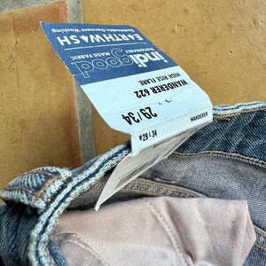 May include: Close-up of blue denim jeans with a high-rise flare. A tag is attached, displaying the brand "indigood" and the style "Wanderer 622". The tag also indicates a size of 29/34. The jeans are made with sustainable fabric and garment washing.