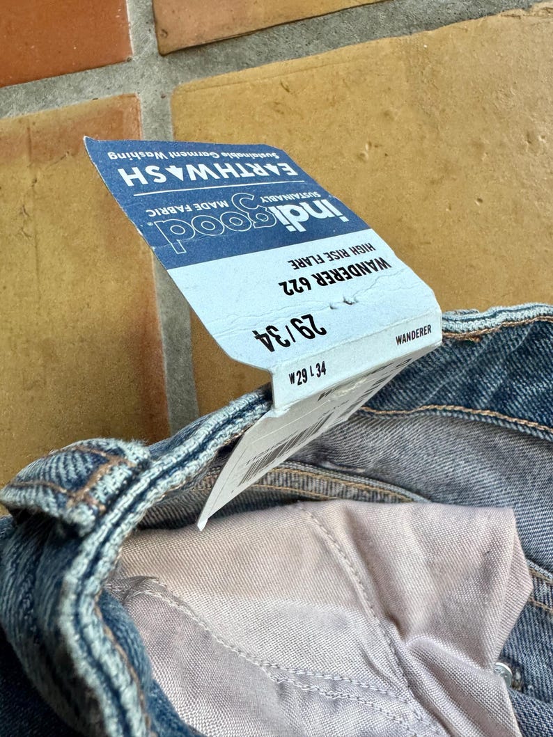 May include: Close-up of blue denim jeans with a high-rise flare. A tag is attached, displaying the brand "indigood" and the style "Wanderer 622". The tag also indicates a size of 29/34. The jeans are made with sustainable fabric and garment washing.