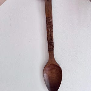 May include: A long, dark brown wooden spoon featuring a carved handle with intricate geometric patterns and a small hole at the top for hanging. The spoon has a smooth, oval-shaped bowl and a straight, textured handle with detailed carvings.