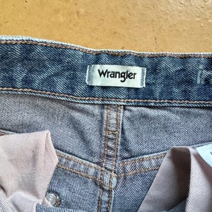 May include: Close-up of blue denim jeans with the brand name "Wrangler" on a white label. The jeans have a classic design with visible stitching and a button closure. The fabric appears to be pre-washed.