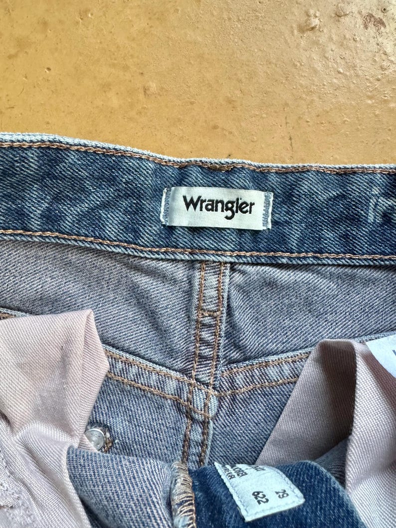 May include: Close-up of blue denim jeans with the brand name "Wrangler" on a white label. The jeans have a classic design with visible stitching and a button closure. The fabric appears to be pre-washed.
