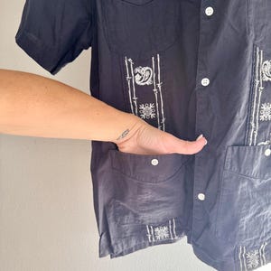 May include: Navy blue short-sleeve shirt with white embroidered details. The shirt features a button-down front, two chest pockets, and decorative embroidery on the front and sleeves. The shirt is made of a lightweight fabric.