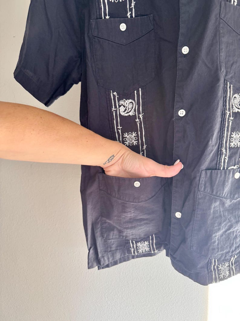May include: Navy blue short-sleeve shirt with white embroidered details. The shirt features a button-down front, two chest pockets, and decorative embroidery on the front and sleeves. The shirt is made of a lightweight fabric.