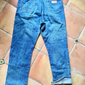 May include: Back view of blue denim jeans with a classic five-pocket design. The jeans have a light wash and a straight leg fit. The Wrangler brand label is visible on the back pocket.