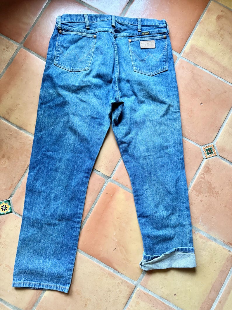 May include: Back view of blue denim jeans with a classic five-pocket design. The jeans have a light wash and a straight leg fit. The Wrangler brand label is visible on the back pocket.