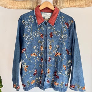May include: A denim jacket with floral embroidery. The jacket features a red collar and is adorned with embroidered flowers in shades of red, orange, and green. The jacket is buttoned up and hangs on a wooden hanger.