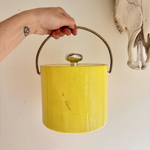 May include: A bright yellow ice bucket with a silver knob on the lid and a gold-colored handle. The cylindrical bucket is held by a hand. A white skull is in the background.