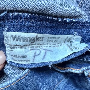 Puede incluir: Unos vaqueros de mezclilla azul con una etiqueta interior blanca que muestra el logotipo de Wrangler, el texto 13MWZPW, 36 x 34, Made in USA, 100% Cotton y las letras manuscritas PT. La tela tiene un tejido visible y costuras de hilo grueso.