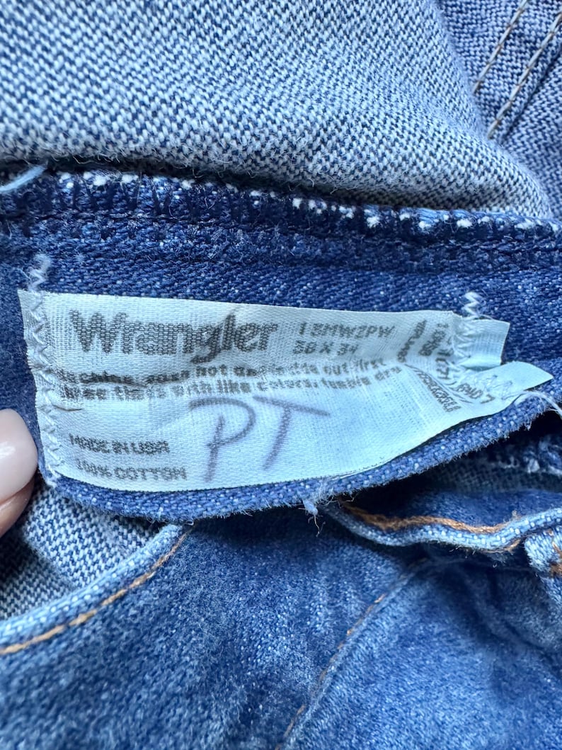Puede incluir: Unos vaqueros de mezclilla azul con una etiqueta interior blanca que muestra el logotipo de Wrangler, el texto 13MWZPW, 36 x 34, Made in USA, 100% Cotton y las letras manuscritas PT. La tela tiene un tejido visible y costuras de hilo grueso.