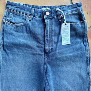 May include: Close-up of blue denim jeans with a button closure and copper rivets. The jeans have front pockets and a tag that reads "NATURAL DYE" and "Wrangler". The jeans are made of a sturdy denim material.