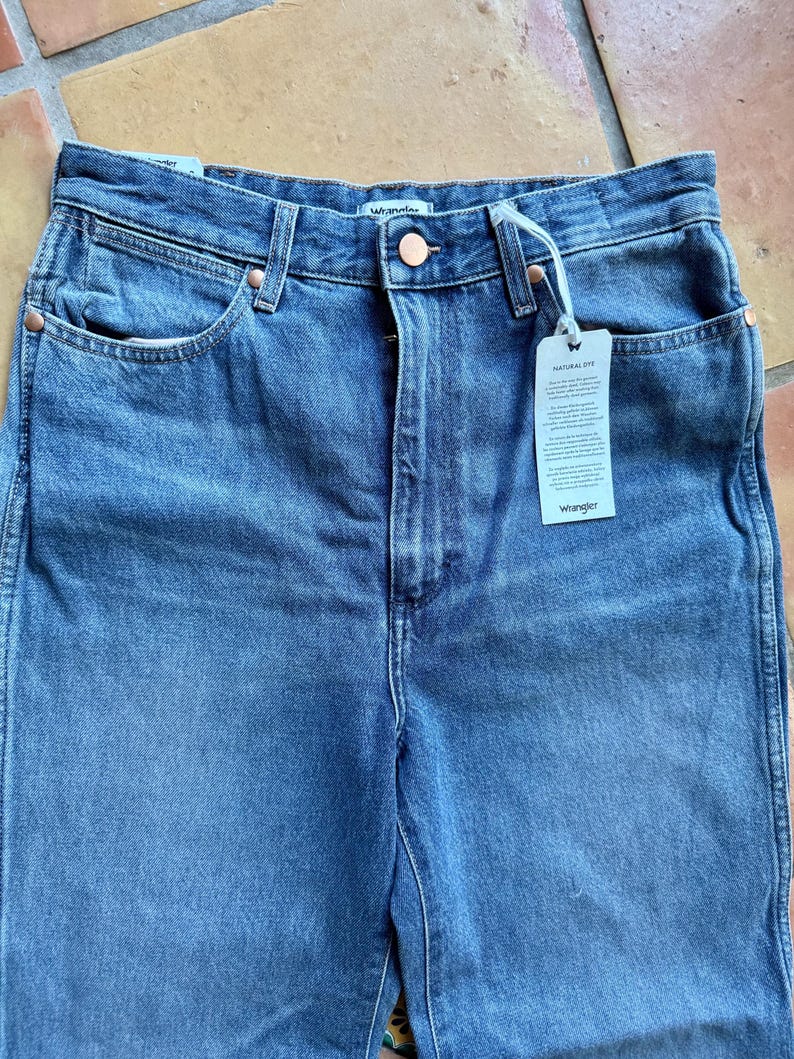 May include: Close-up of blue denim jeans with a button closure and copper rivets. The jeans have front pockets and a tag that reads "NATURAL DYE" and "Wrangler". The jeans are made of a sturdy denim material.
