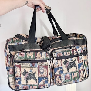 May include: A tapestry-style travel bag with a repeating pattern of dogs, houses, and other imagery. The bag has two zippered front pockets, black handles, and a shoulder strap. The bag is held up by a hand.