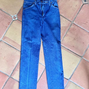 May include: Blue denim jeans with a zipper fly and classic five-pocket design. The jeans are a medium wash and appear to be vintage, with a straight leg cut. The jeans are laid flat on a tiled surface.