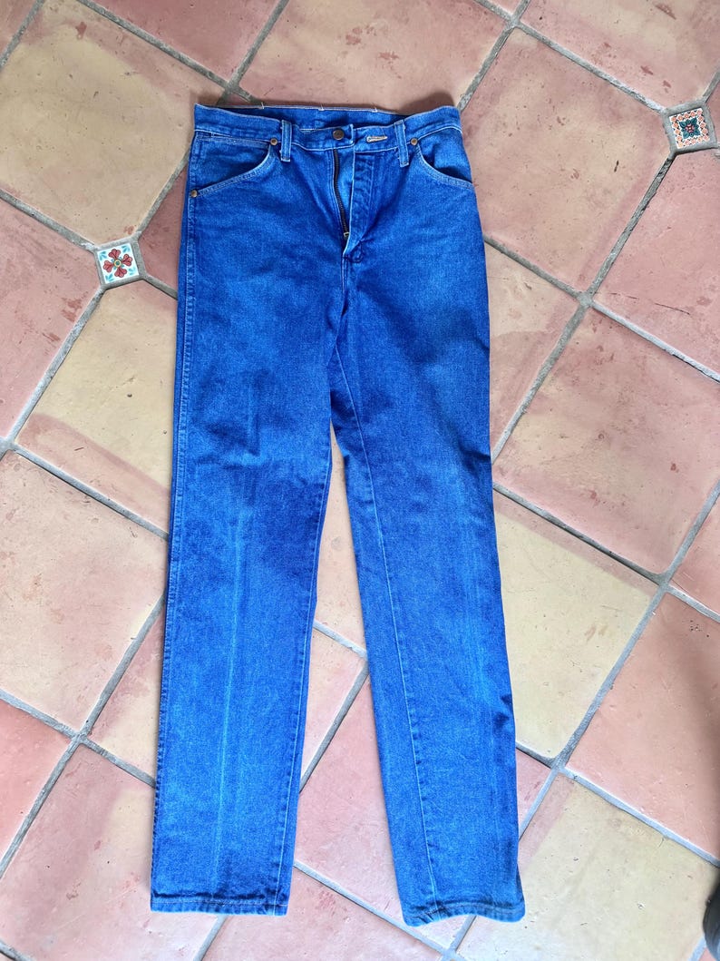 May include: Blue denim jeans with a zipper fly and classic five-pocket design. The jeans are a medium wash and appear to be vintage, with a straight leg cut. The jeans are laid flat on a tiled surface.