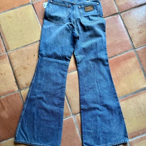 May include: Vintage blue denim Wrangler jeans with a flared leg. The jeans have classic five-pocket styling, a Wrangler leather patch, and a tag attached. The jeans are laid flat on a tiled surface.