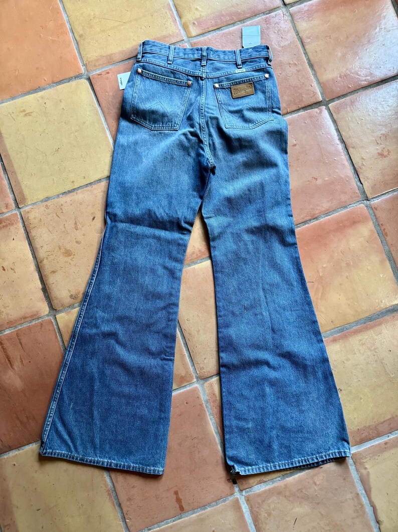 May include: Vintage blue denim Wrangler jeans with a flared leg. The jeans have classic five-pocket styling, a Wrangler leather patch, and a tag attached. The jeans are laid flat on a tiled surface.