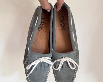 May include: A pair of gray suede moccasin loafers with white stitching and fringe detail. The shoes have white laces and the brand name "Minnetonka Moccasin" is visible on the insole.