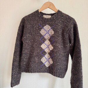 May include: A dark gray argyle sweater with a crew neck and long sleeves. The argyle pattern features light beige and purple diamond shapes. The sweater is hanging on a wooden hanger against a white wall.