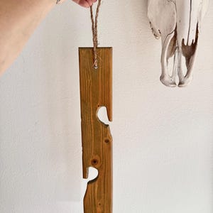 May include: A rustic, wooden wine bottle holder with heart-shaped cutouts. The holder is stained a warm brown and suspended by a natural rope. The design is simple and would complement a farmhouse or rustic decor style.