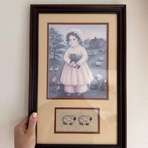 May include: Framed artwork featuring a young girl in a pink dress holding a black cat, set in a pastoral scene with sheep and a farmhouse. Below, two embroidered sheep are displayed.