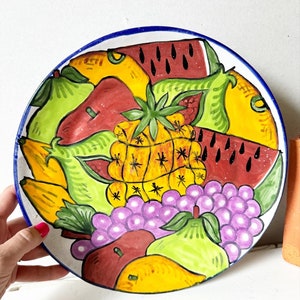 May include: A white ceramic plate with a blue rim, hand-painted with a colorful assortment of fruit, including watermelon, pineapple, grapes, pears, and bananas.