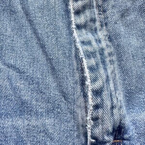 May include: Close-up of light blue denim fabric. The image highlights the texture and stitching details of the denim material, with a focus on the seam and the overall weave of the fabric.