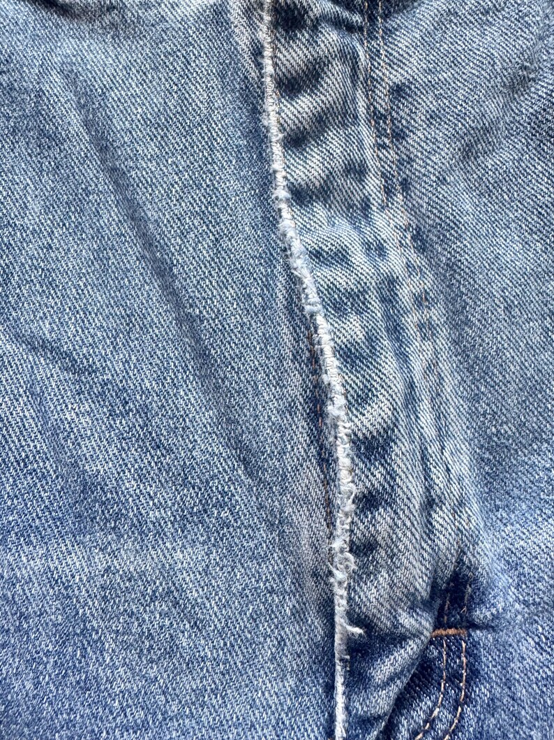 May include: Close-up of light blue denim fabric. The image highlights the texture and stitching details of the denim material, with a focus on the seam and the overall weave of the fabric.