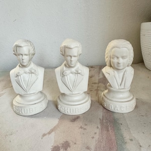 May include: Three white ceramic busts of composers. Two are labeled "SCHUBERT" and one is labeled "BACH". The busts feature detailed facial features, formal attire, and are set on round bases. The background is a neutral color.