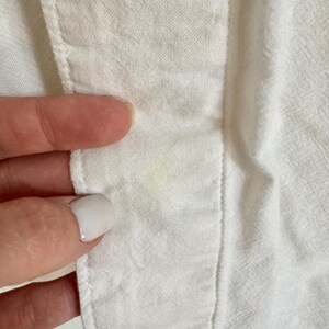 May include: Close-up of white fabric with visible stitching and a seam. The fabric appears to be a lightweight material, possibly cotton or linen. A finger is touching the fabric, highlighting the texture and construction of the garment.