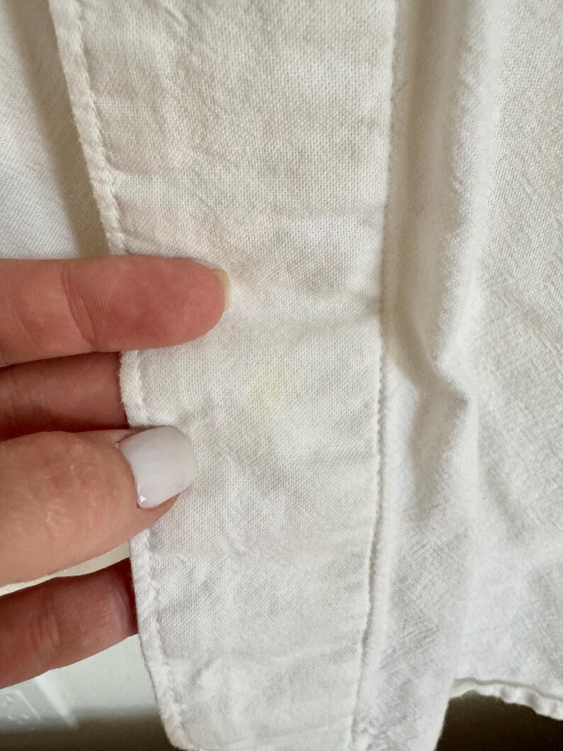 May include: Close-up of white fabric with visible stitching and a seam. The fabric appears to be a lightweight material, possibly cotton or linen. A finger is touching the fabric, highlighting the texture and construction of the garment.