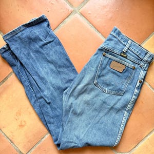 K&ouml;nnte beinhalten: Hellblaue Jeans mit klassischer Passform. Die Jeans hat einen braunen Lederaufn&auml;her auf der Ges&auml;&szlig;tasche mit dem Markennamen "Wrangler". Die Jeans liegen auf einer Terrakotta-Fliesenoberfl&auml;che.