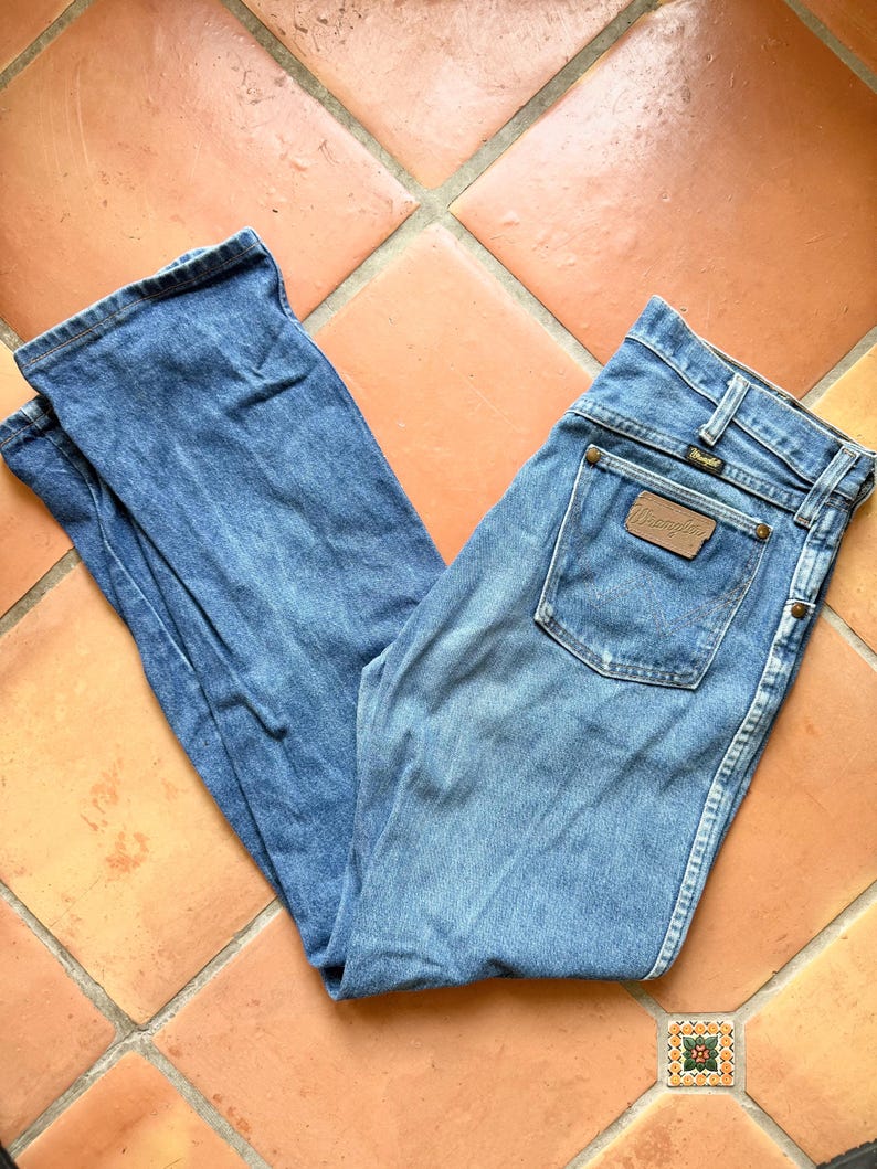 K&ouml;nnte beinhalten: Hellblaue Jeans mit klassischer Passform. Die Jeans hat einen braunen Lederaufn&auml;her auf der Ges&auml;&szlig;tasche mit dem Markennamen "Wrangler". Die Jeans liegen auf einer Terrakotta-Fliesenoberfl&auml;che.