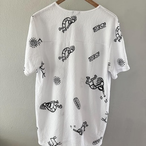 May include: White short-sleeved shirt with a textured surface, adorned with black Kokopelli figures, sun symbols, and other abstract designs. The shirt is hanging on a wooden hanger against a neutral background.
