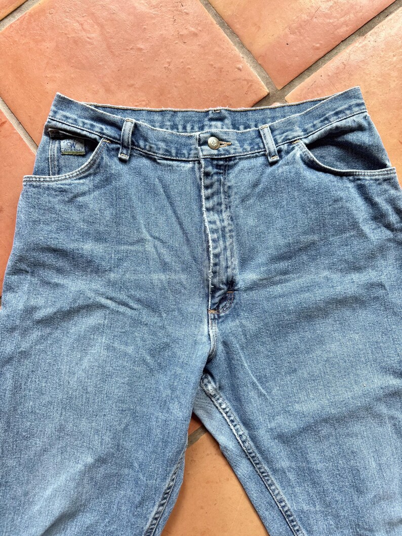 May include: Light blue denim jeans with a button closure and front and back pockets. The jeans have a classic five-pocket design and a straight leg fit. The Calvin Klein logo is visible on the front pocket.