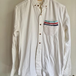 May include: A white button-down shirt with a unique crochet pocket. The pocket features a striped design in red, blue, and turquoise. The shirt has a classic collar and wooden buttons. The brand name "Koto" is visible on the collar.