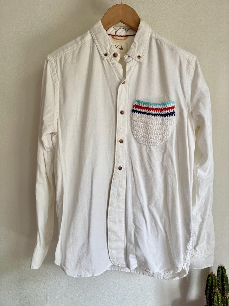 May include: A white button-down shirt with a unique crochet pocket. The pocket features a striped design in red, blue, and turquoise. The shirt has a classic collar and wooden buttons. The brand name "Koto" is visible on the collar.
